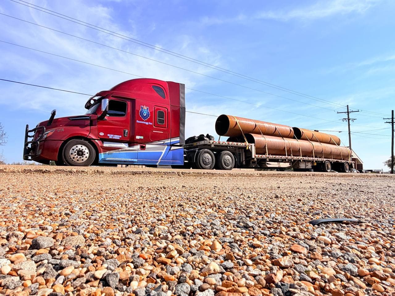 Red Snowmen Express truck hauling large steel pipes on a flatbed trailer, photographed from a dramatic low angle